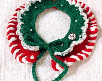 May include: A red and white striped crocheted collar with a green crocheted bow and silver beads. The collar has a green crocheted ruffle with silver beads and a gold charm with the letter "M" on it.