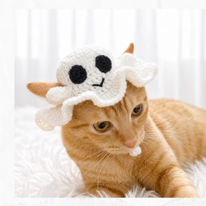 Puede incluir: Un gorro de fantasma tejido a ganchillo en color blanco con ojos circulares negros y una pequeña boca negra bordada. El gorro tiene un borde ondulado y con volantes, y lo lleva un gato naranja. El texto Crochet Pattern aparece a la izquierda.