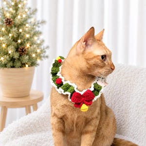 May include: A crocheted pet collar featuring a green ruffled wreath design with white trim, accented by small red pom-poms, a prominent red crocheted bow at the center, and a small yellow bell hanging from the bottom of the bow.
