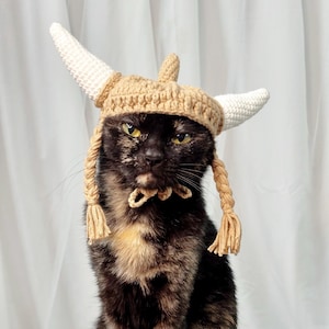 Pode incluir: Um gato tartaruga usando um chapéu viking de crochê feito à mão. O chapéu é bege com dois chifres brancos e detalhes de trança de fio. O gato tem olhos amarelos e uma expressão séria.