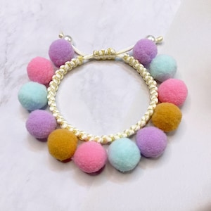 May include: A white string bracelet with colorful pom-poms. The pom-poms are in shades of pink, purple, yellow, and blue.