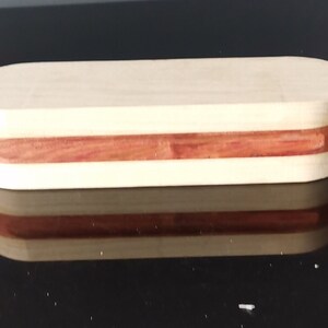 Op de afbeelding: Een houten doos met een lichtbruine bovenkant en een roodbruine inleg. De doos is ovaalvormig en heeft een gladde afwerking.