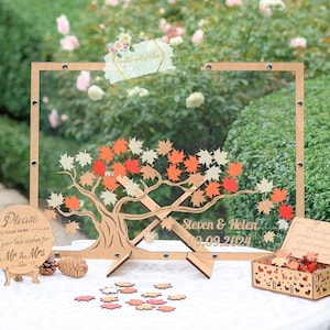 Könnte beinhalten: Ein hölzernes Hochzeitsgästebuch mit einem klaren Acrylrahmen. Der Rahmen zeigt ein lasergeschnittenes Design eines Baumes mit Blättern in Orange-, Rot- und Gelbtönen. Die Blätter sind dazu gedacht, entfernt und von den Gästen signiert zu werden. Der Text "Steven & Helen, 09.09.2024" ist in den Rahmen graviert.