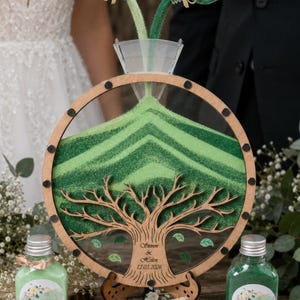 May include: A circular wooden sand ceremony frame with a tree cutout, featuring the engraved text Steven & Helen 12.03.2026. Two glass bottles pour light and dark green sand into the frame through a clear funnel, creating layered patterns.