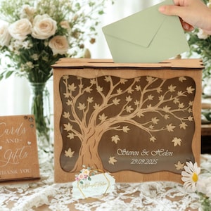 以下が含まれることがあります： 名前と日付が刻まれた、木の模様の木製ウェディングカードボックス。 手が明るい緑色の封筒をスロットに入れています。 小さな木製の看板には「カードとギフト」と書かれています。