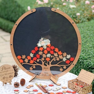 May include: A round wooden guest book with a clear acrylic cover, featuring a fall-themed tree with red, orange, and brown leaves. The leaves are attached with pins and can be removed by guests. The guest book also includes a small wooden box, two wooden pens, and a sign that says "Please leave your name and your best wishes for Mr. & Mrs." The sign also includes the names "Steven & Helen" and the date "09.09.2023".