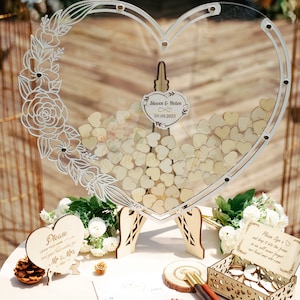Può includere: Un libro degli ospiti a forma di cuore in acrilico trasparente con un design floreale e pezzi a forma di cuore in legno all'interno. Sono visualizzati i nomi "Steven & Helen" e la data "09.09.2023". Sono presenti anche cartelli in legno aggiuntivi e una scatola con pezzi a forma di cuore.