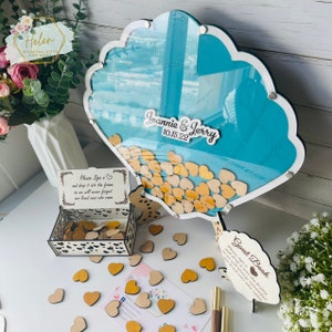 May include: A teal seashell-shaped guest book with a clear acrylic front and a wooden base. The guest book is filled with small wooden hearts. The text "Joannie & Jerry 10.15.22" is on the front of the guest book.