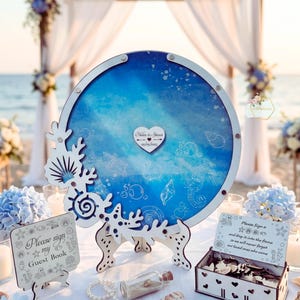 May include: A circular guest book alternative with a blue ocean design and white coral detailing. The centre has a heart-shaped tag with names and a date. Additional decor includes a sign that says "Please sign my Guest Book.".
