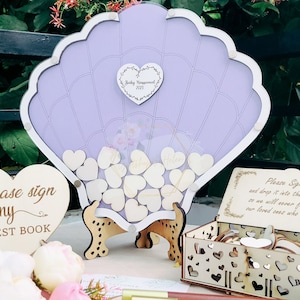 Könnte beinhalten: Ein violettes Muschel-förmiges Gästebuch mit einem Holzständer. Das Gästebuch ist gefüllt mit kleinen Holzh Herzen, die Gäste signieren und in die Box legen können. Die Box ist aus Holz und hat ein herzförmiges Design. Das Schild sagt "Bitte unterschreiben Sie mein Gästebuch".