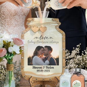 Peut inclure: Un cadre de cérémonie de sable en forme de bocal en bois avec une photo centrale d'un couple, portant le texte love today tomorrow always et Steven & Helen 09.09.2025. Le cadre est soutenu par un support en bois et inclut deux bouteilles.