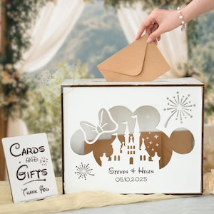 May include: A white wooden card box with a Mickey Mouse silhouette design, a bow, castle, and fireworks cutouts. The box has the names "Steven & Helen" and the date "05.10.2025" engraved. A person is placing a brown envelope into the slot.