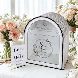 May include: A white wedding post box with a clear front panel, arched top, and black trim. The box features the text "WEDDING POST" and a circular design with initials and a date. A small sign reads "Cards and Gifts".