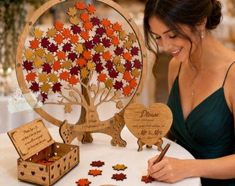 May include: A wooden wedding guest book alternative featuring a tree design with colorful leaf cutouts. Includes a heart-shaped sign, a box, and a pen for guests to sign. The tree is surrounded by loose leaf cutouts.