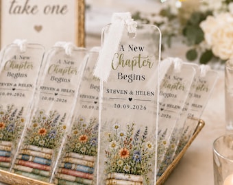 May include: A clear acrylic rectangular bookmark featuring a white ribbon at the top. The text reads A New Chapter Begins, STEVEN & HELEN, and 10.09.2026. Below the text is a colorful illustration of a stack of books topped with wildflowers.