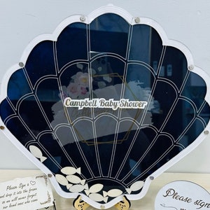 May include: A white seashell-shaped frame with a black and white design. The frame is filled with white fish shapes and has a sign that says "Campbell Baby Shower".