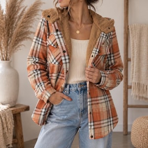 May include: A long-sleeve button-up jacket featuring an orange, white, and black plaid pattern. The jacket includes a tan sherpa-lined hood, a front button closure, and two chest pockets with buttoned flaps, worn over a white ribbed tank top and jeans.