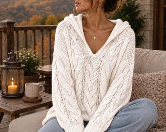 May include: An off-white cable knit hooded jumper with a V-neckline and long sleeves, paired with light blue denim jeans. The jumper features a prominent vertical braided pattern throughout the front and ribbed cuffs at the wrists.