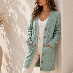 May include: A sage green cable-knit cardigan featuring a V-neckline, long sleeves, and a button-down front with dark brown buttons. The garment includes two front patch pockets and ribbed cuffs and hem, layered over a white top and light pants.