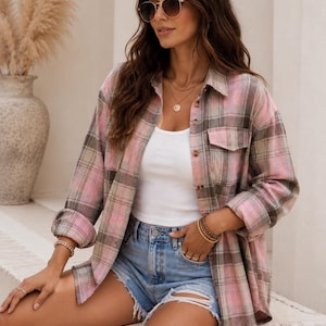 May include: A long-sleeve button-down shirt featuring a pink, beige, and brown plaid pattern with two chest pockets. The shirt is worn over a plain white tank top paired with light blue denim shorts that have a distressed, frayed hemline.