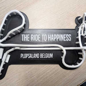 May include: A black and white decorative item featuring the text "THE RIDE TO HAPPINESS" and "PLOPSALAND BELGIUM". The item has a roller coaster design with white tracks and a black base.
