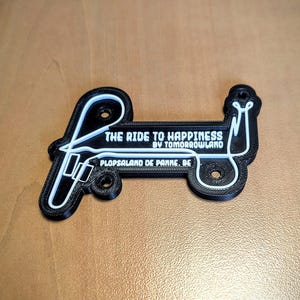 Peut inclure: Un porte-clés imprimé en 3D en noir et blanc avec le texte "THE RIDE TO HAPPINESS BY TOMORROWLAND" et "PLOPSALAND DE PANNE. BE". Le porte-clés a un design stylisé unique.
