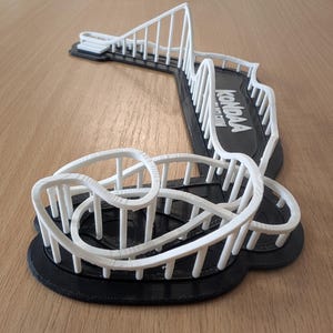 Puede incluir: Modelo impreso en 3D de una montaña rusa con vías y soportes blancos sobre una base negra. El modelo incluye un bucle, una caída y el texto "Kondaa". La montaña rusa está sobre una superficie marrón claro.