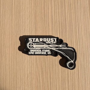 Puede incluir: Llavero impreso en 3D en blanco y negro con el logotipo de Stardust Racers, una pista de montaña rusa y el texto "Universal Studios Epic Universe, USA".