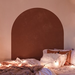 May include: A bedroom with a white bed with a brown faux fur throw blanket and white pillows. The wall behind the bed has a large brown arch painted on it. There are string lights draped over the bed and a small table with candles on it.