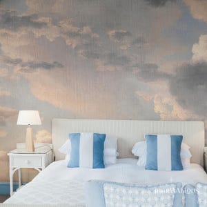 May include: A bedroom scene featuring a bed with white bedding and two blue and white striped pillows. A white nightstand with a lamp sits to the left. The wall behind the bed is painted with a cloudy sky mural.