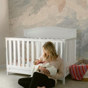 May include: A woman sits on a white carpet holding a newborn baby in a white blanket. The baby is wearing a burgundy headband. The woman is wearing black pants and a light brown shirt. A white crib with a white and pink patterned sheet is in the background. The wall behind the crib is painted with a light blue and pink watercolor design. The text "marWALlous" is in the bottom right corner of the image.