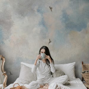 May include: A person sitting on a bed, holding a light blue mug. The background features a mural with a sky-like design in shades of blue, white, and gray. The person is wearing a light gray pajama set. The word "marWALious" is visible.