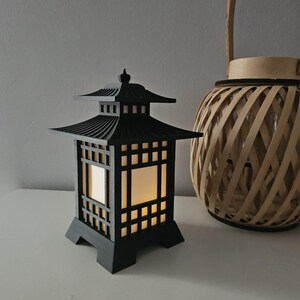 Pode incluir: Uma lanterna de madeira preta com um design tradicional japonês. A lanterna tem uma base quadrada e um telhado em camadas. A lanterna é iluminada por dentro, projetando um brilho quente.