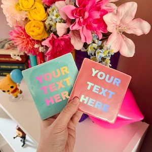 Op de afbeelding: Twee vierkante keramische onderzetters met een glanzende afwerking. De ene is lichtblauw met felroze tekst die YOUR TEXT HERE leest. De andere is zachtroze met lichtblauwe tekst die YOUR TEXT HERE leest. Beide items worden samen vastgehouden.