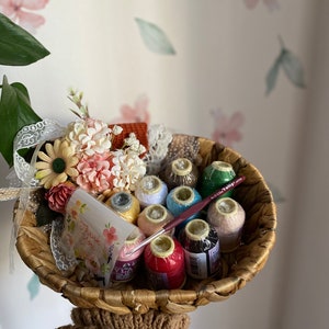 Peut inclure: Un panier en osier rempli de bobines de fil de couleurs et d'un crochet. Le panier est décoré d'un petit bouquet de fleurs et d'une carte qui dit "Merci".
