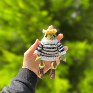 Puede incluir: Una pequeña rana de punto verde que lleva un jersey a rayas blancas y negras, un sombrero amarillo y botas marrones. La rana está siendo sostenida en una mano contra un fondo verde borroso.