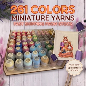 May include: A collection of miniature yarn spools in a wooden organiser tray, featuring 261 colours. A beige drawstring pouch with a printed illustration of three dolls and the text Spicaartdoll is included as a gift, alongside loose spools.