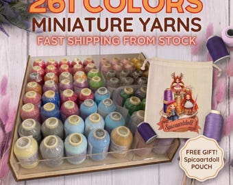 May include: A collection of miniature yarn spools in a wooden organizer tray, featuring 261 colors. A beige drawstring pouch with a printed illustration of three dolls and the text Spicaartdoll is included as a gift, alongside loose spools.