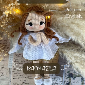 Pode incluir: Padrão de crochê para uma boneca anjo branca com cabelo castanho, vestindo um vestido branco com detalhes dourados. O padrão é chamado de "Ramaela" e está escrito em inglês.