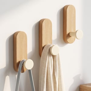 May include: Three wall-mounted hooks featuring vertical light-toned wooden backplates with rounded ends and protruding cream-colored cylindrical pegs. One hook holds a grey strap, another holds a beige fabric item, and one remains empty.