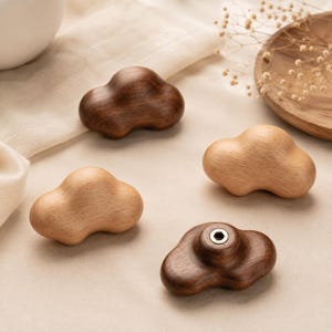 Puede incluir: Cuatro pomos para armario con forma de nube fabricados en madera maciza. Dos pomos presentan un acabado marrón oscuro, mientras que los otros dos tienen un tono de madera natural más claro. Cada pomo tiene una superficie lisa y redondeada.