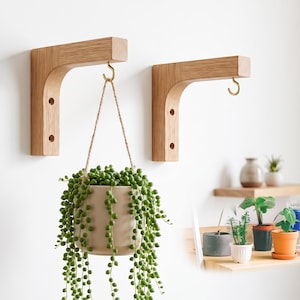 Può includere: Due supporti a parete in legno chiaro dal design curvo, ciascuno dotato di un gancio in metallo color ottone. Un supporto sostiene un vaso in ceramica beige contenente una pianta di senecio rowleyanus con perle verdi.