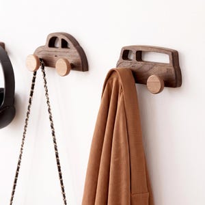 May include: Two dark brown wooden wall hooks shaped like vehicles. One hook is shaped like a sedan and holds a black chain strap, while the other is shaped like a van and holds a brown garment. Each hook features round light wood wheel accents.