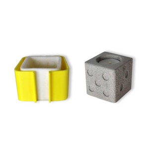 May include: Two decorative items: a yellow and white square container and a gray concrete cube with a candle inside. The container has a square opening and a white interior. The cube has curved indentations on its sides.