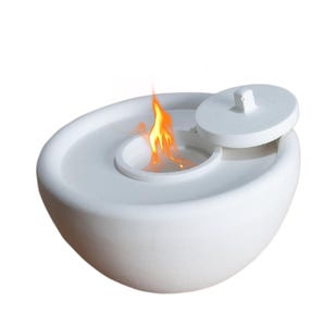 May include: White ceramic fire pit with a lid partially open, revealing a small flame inside. The fire pit is round and has a smooth, slightly textured surface.