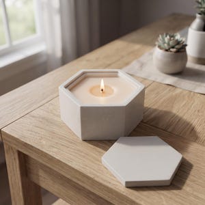 Peut inclure: Un bougeoir hexagonal en béton gris clair avec une bougie allumée à l'intérieur, présentant une finition mate lisse et un couvercle hexagonal amovible assorti posé à côté sur une surface en bois, formant un objet de décoration fonctionnel.