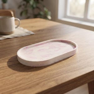 May include: An oval-shaped tray featuring a smooth, matte finish with a marbled pattern of soft pink and white swirls. The tray has a raised edge and a flat base, constructed from a solid, stone-like material with a subtle speckled texture.