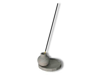 May include: A gray concrete incense holder with a round base and a small, round, gray vase on top. A black incense stick is sticking straight up from the vase.