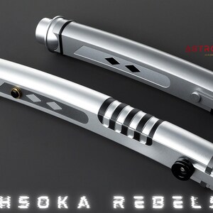 May include: Two silver lightsabers with black and gold accents, featuring a diamond pattern and black stripes. The text "AHSOKA REBELS" is displayed at the bottom. The logo "ASTROMX SABERS" is in the upper right corner.