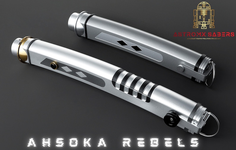May include: Two silver lightsabers with black and gold accents, featuring a diamond pattern and black stripes. The text "AHSOKA REBELS" is displayed at the bottom. The logo "ASTROMX SABERS" is in the upper right corner.